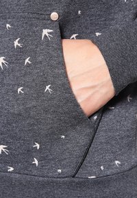Sweat-shirt gris avec motifs d'oiseaux blancs ; doté d'une poche avant et d'un petit accent métallique. Le tissu a une texture lisse et douce.