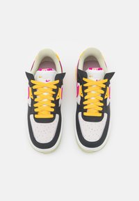 Zapatillas deportivas en blanco y negro con cordones amarillos, detalles en rosa y puntera perforada. Cuentan con suela de goma y cuello acolchado.