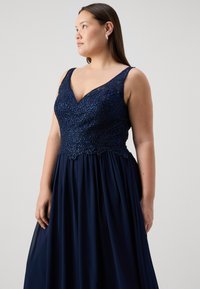 Mornarska obleka z beaded korzetom in V-izrezom ter širokimi naramnicami; padajoča krila iz lahkega materiala, ki ustvarjajo elegantne drapirne učinke.