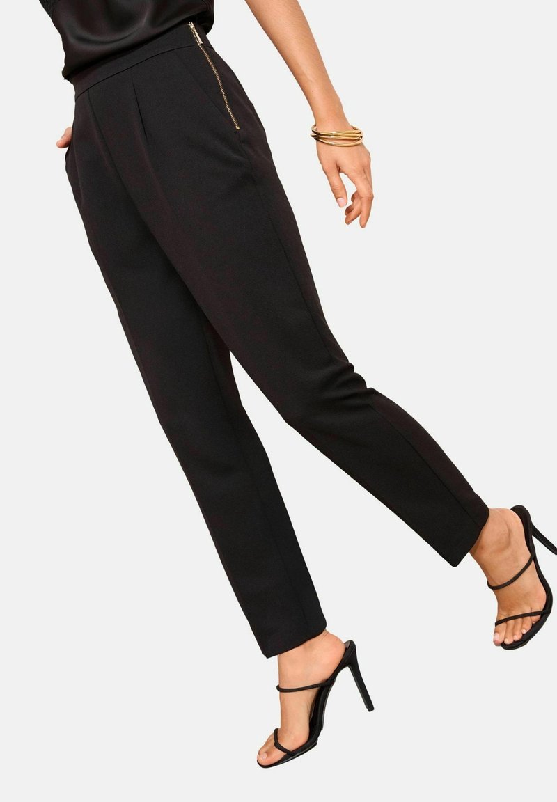 Femme portant un pantalon noir fuselé avec des détails de fermeture éclair dorée et des sandales à talons hauts noires à brides, montrant les jambes et un bras avec des bracelets dorés.