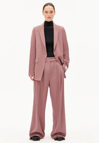 Maßgeschneiderter pinker Anzug mit weiten Hosen und einem passenden Blazer, kombiniert mit einem schwarzen gerippten Rollkragenpullover und schwarzen Schuhen mit Akzenten.