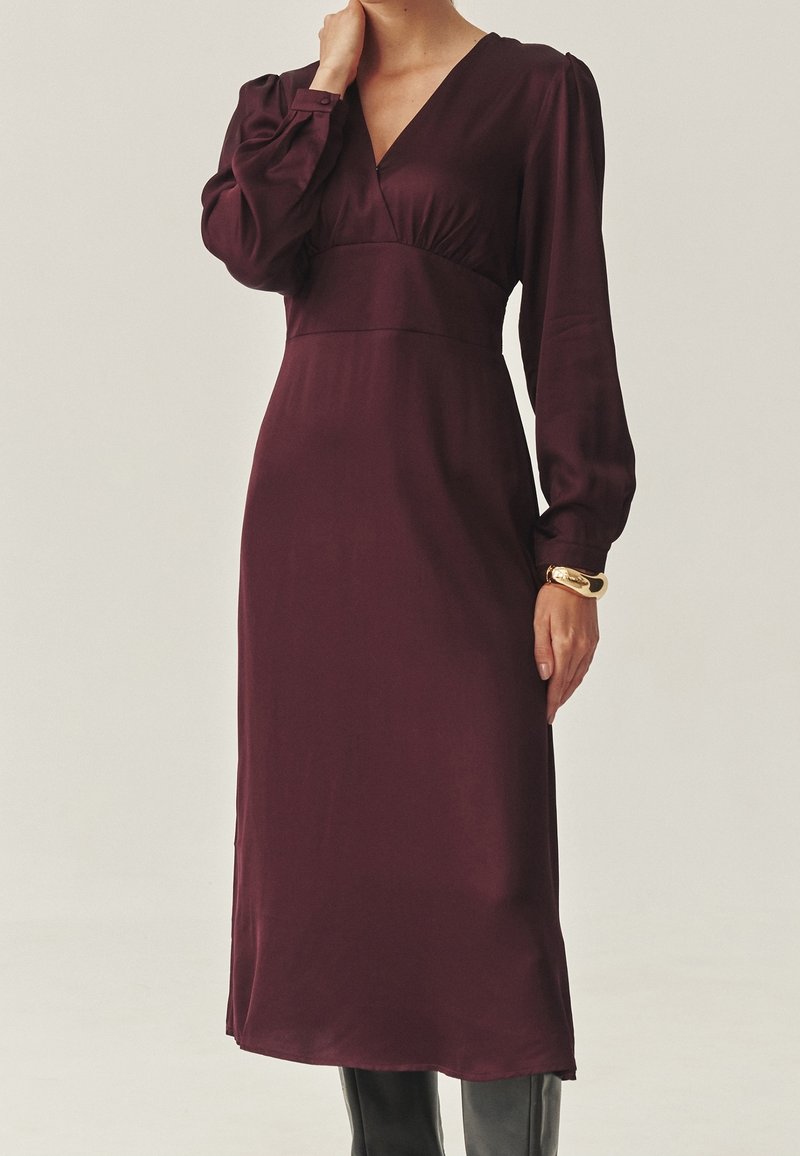 Femme portant une robe midi à manches longues, de couleur violet foncé, avec un col en V et un bracelet en or au poignet gauche.