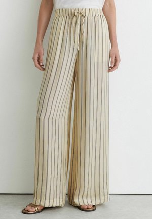 Beige weite Hosen mit vertikalen schwarzen Streifen, elastischem Bund mit Kordelzug, getragen mit flachen braunen Sandalen und einem weißen Top.