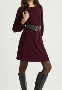 Robe bordeaux à manches longues avec une ceinture noire texturée agrémentée d'accents colorés. La modèle porte des bottes noires à motif reptile montant jusqu'aux genoux.