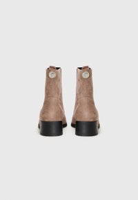 Botines de ante marrón con un tacón grueso negro, que presentan acentos metálicos redondos en la parte trasera y una textura suave.