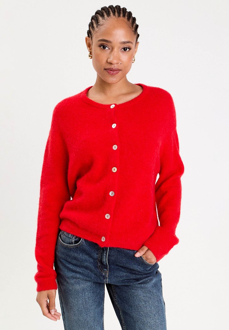 Kvinde med fletninger, iført en rød knap cardigan og blå jeans, stående med den ene hånd i lommen foran en ensfarvet baggrund.