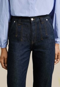 Donkerblauwe denim jeans met een hoge taille, voorzien van oranje stiksels, een knoopsluiting en voorzakken. Gecombineerd met een lichtblauwe gestreepte blouse.