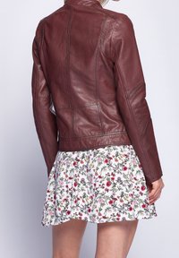 Mujer con una chaqueta de cuero marrón ajustada sobre una falda plisada blanca con flores, de pie contra un fondo claro y liso.