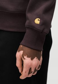 Καφέ φούτερ με ριμπ μανσέτες. Χρυσό λογότυπο στη μ ανσέτα. Το χέρι έχει κηλιώδη, σκούρο καφέ τόνο δέρματος. Μαύρο φόντο.