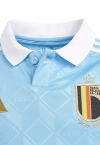 Ljusblå pikétröja med vit krage, knäppning och ett texturerat mönster. Inkluderar ett broderat emblem för Belgien nationallag.