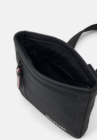 Borsa a tracolla nera in materiale sintetico con superficie testurizzata, chiusura con zip e dettagli rossi, bianchi e blu. L'interno è foderato in tessuto nero.