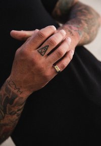 Juodas žiedas su stačiakampiu aukso akcentu, dėvimas ant rankos su tatuiruotėmis. Matoma odos spalva, dėmesys skiriamas žiedo ir rankos detalėms.