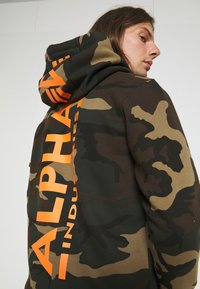 Zöld és barna árnyalatú álcázó kapucnis pulóver narancssárga "ALPHA INDUSTRIES" felirattal a háton. Csatlakoztatható kapucni és puha anyag jellemzi.