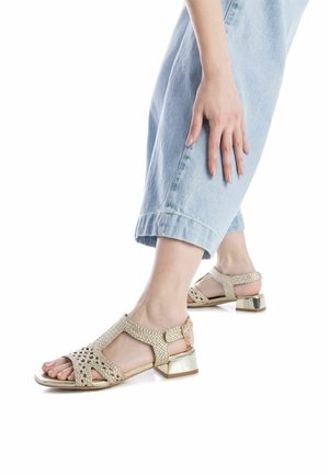 Sandali in oro intrecciato con design a ritaglio, un tacco basso a blocco e cinturino alla caviglia, abbinati a jeans in denim blu chiaro.