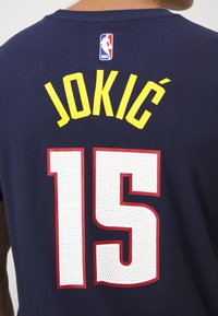 Maillot NBA bleu marine avec le nom jaune « Jokić » et le grand numéro blanc 15 bordé de rouge au dos.