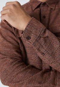 Mees, kellel on seljas pruun tekstureeritud villane särk, millel on nööridega mansetid, käsi tõstetud ja rinnale ristatud, näidates kangast ja nööpe.