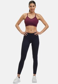 Bordeaux sportbeha met dunne banden, gecombineerd met hoge marineblauwe leggings met subtiele stiksel-accenten. Draag met witte sportschoenen.