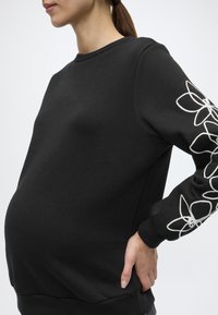 Μαύρο φούτερ με μακριά μανίκια και στρογγυλεμένο λαιμόκοψη. Διακοσμημένο με λευκό κεντητό λουλούδι κατά μήκος του αριστερού μανικιού. Μαλακό υλικό, άνετη εφαρμογή.