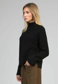 Femme blonde portant un pull à col roulé noir côtelé à manches longues et un pantalon marron avec de grandes poches avant, se tenant de profil.