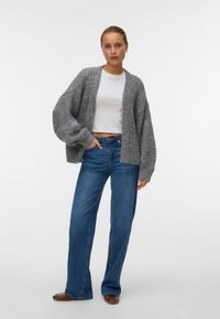 Γυναίκα που φοράει γκρι μάλλινη ζακέτα με χοντρή πλέξη, λευκό κοντό τοπ, μπλε τζιν με φαρδιά μπατζάκια και καφέ παπούτσια, στέκεται μπροστά σε απλό λευκό φόντο.