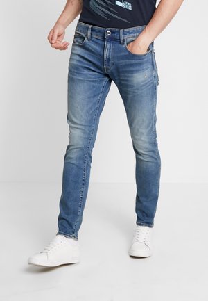 Mann trägt schlank geschnittene blaue Jeans und weiße Sneaker, eine Hand in der Tasche, steht vor einem schlichten hellen Hintergrund.