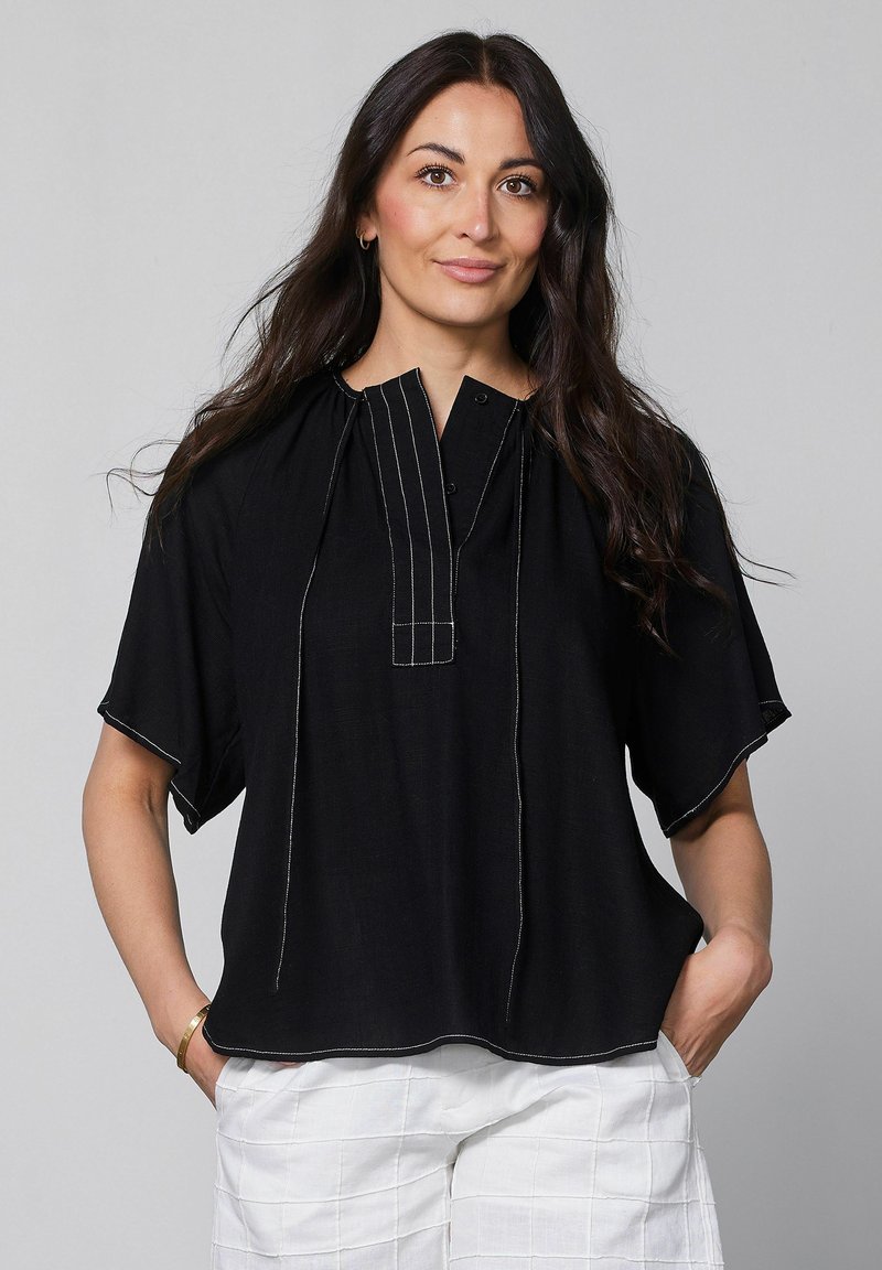 Femme aux longs cheveux foncés portant une chemise noire ample avec des coutures blanches et un pantalon blanc, debout les mains dans les poches.