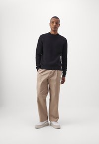 Svart strikkegenser, beige skreddersydde bukser og hvite sneakers. Enkel design med ribbestrikkede mansjetter og rette bukser, laget av mykt stoff.