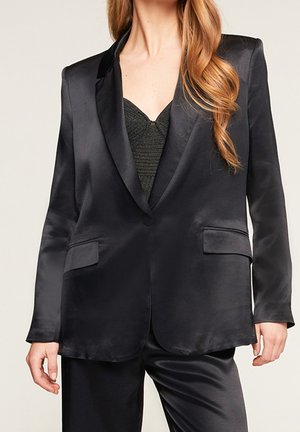 Woman wearing a black satin blazer over a textured black top, paired with matching black satin pants, standing against a plain background.