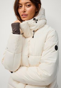 Veste rembourrée de couleur crème avec un col montant, quincaillerie noire et poignets élastiques. Présente des motifs cousus et un patch logo sur la manche.