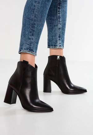 Paire de bottines en cuir noir avec talons carrés et fermetures éclair sur les côtés, portées avec un jean en denim bleu coupé au-dessus des chevilles.