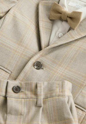 Costume en tartan beige avec une texture douce, doté d'un nœud papillon, de deux boutons et de coutures détaillées, mettant en valeur des accents dorés et orangés.