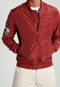 Czerwona nylonowa kurtka bomberka z ściągaczami przy mankietach i u dołu, z naszywką na lewym rękawie oraz zamkiem z przodu. Gładka tekstura.