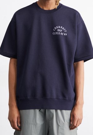 Personne portant un sweat-shirt bleu marine à manches courtes surdimensionné avec le texte "Carhartt MIP Class of 89" sur la poitrine et un pantalon gris clair.