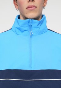 Ανοιχτό μπλε ζακέτα με φερμουάρ, που διαθέτει ψηλό γιακά, ναυτικό οριζόντιο σχέδιο στο κάτω μέρος και λευκή διακοσμητική γραμμή. Λείος υφαντικός ιστός.