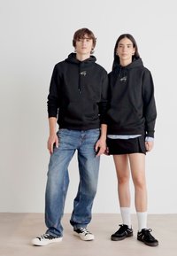Čierne mikiny s prednými klokankami, okrúhlymi výstrihmi a šnúrkami. Džínsy na mužskom modeli a sukňa na ženskému modeli.