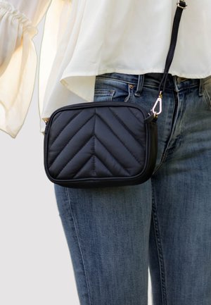 Black quilted shoulder bag with a chevron pattern, featuring a smooth texture, gold hardware, and an adjustable strap, worn with denim jeans.