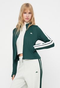 Zeleni sportski zip-up jakna s bijelim i mornarsko plavim prugama na rukavima, napravljena od rastezljivog materijala. Ima mali logotip na prsima i prilagođeni dizajn.