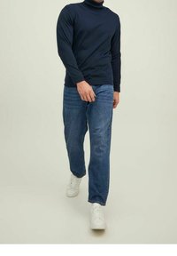 Maglione a collo alto blu navy con maniche lunghe, abbinato a jeans azzurri e sneakers bianche, che mettono in risalto una silhouette casual e aderente.