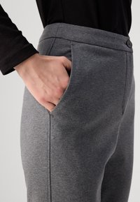 Grå joggebukser med en glatt tekstur, som har sidelommer og knappelukking i midjen, vist med en hånd i lommen.