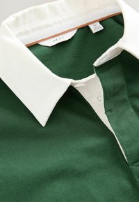 Cămașă polo verde cu guler alb, având închidere cu nasturi și textură ribbed. Eticheta este vizibilă în zona gulerului.