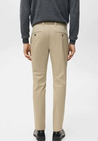 Mężczyzna ubrany w beżowe, dopasowane spodnie z guzikami na tylnych kieszeniach, ciemną koszulę z długim rękawem oraz czarne eleganckie buty, stojący na jednolitym tle.