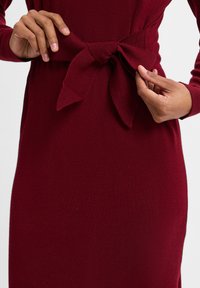 Bordeauxfarbenes Kleid aus strukturiertem Stoff, mit einem Taillengürtel in Schleifenform, Hände, die die Schleife anpassen, Nahaufnahme der Details.