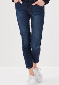 Jean en denim bleu foncé à coupe ajustée, avec un léger délavage, des surpiqûres contrastées et une longueur chevilles raccourcie, associés à des baskets blanches.