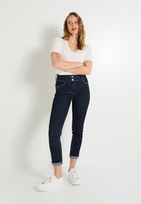 Jeans de mezclilla oscura de cintura alta con un corte tapered, dos grandes botones en la parte delantera y puños doblados. Combinados con una camiseta blanca de manga corta y zapatillas.