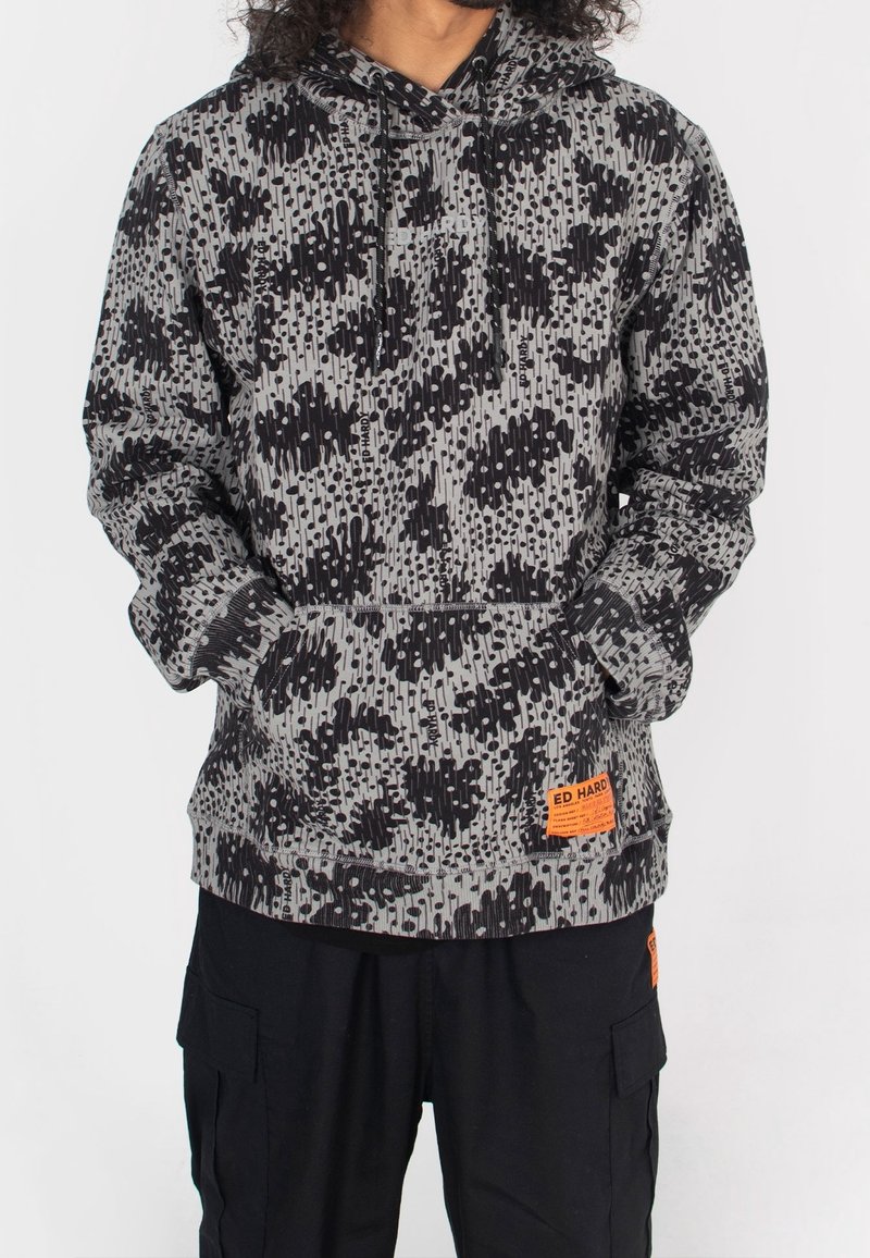 Personne portant un sweat à capuche gris et noir tacheté avec une poche avant et un patch orange, associé à un pantalon cargo noir avec des poches latérales.