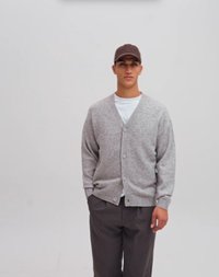 Jonge man die een grijs vest, wit T-shirt, donkere broek en een bruine pet draagt, staand tegen een eenvoudige lichte achtergrond.