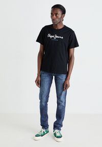 Sort bomulds T-shirt med hvid "Pepe Jeans London" logo, parret med blå denim bukser og grønne og hvide sneakers, stående på en neutral baggrund.