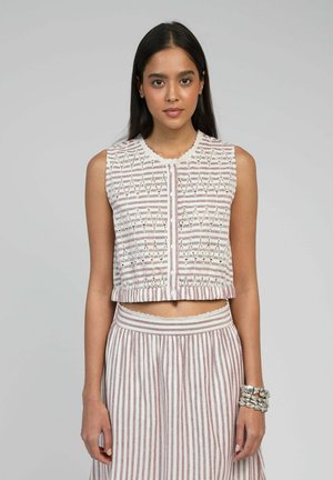 Jeune femme portant un haut court sans manches boutonné avec un motif en zigzag et une jupe taille haute à rayures verticales, bracelets argentés au poignet droit.