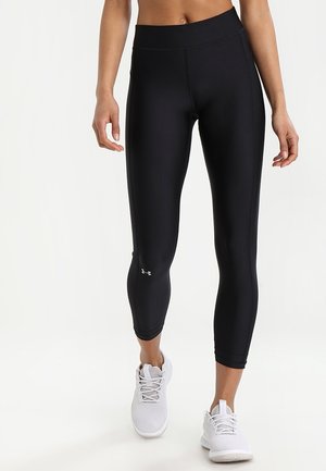 Kobieta nosząca czarne krótkie legginsy z logo Under Armour przy łydce, w połączeniu z białymi sportowymi butami, stojąca na białym tle.