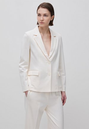 Woman wearing an off-white tailored blazer with two buttons and matching trousers, standing against a plain light grey background.
